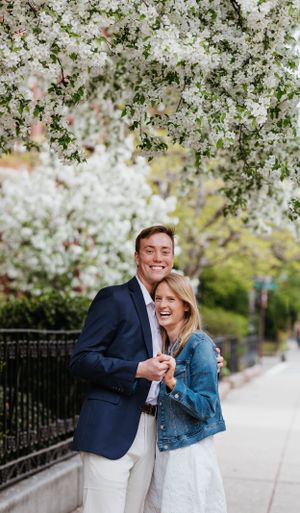 Happy couple embracing under white spring blossoms on a city sidewalk, man in navy blazer and woman in denim jacket.