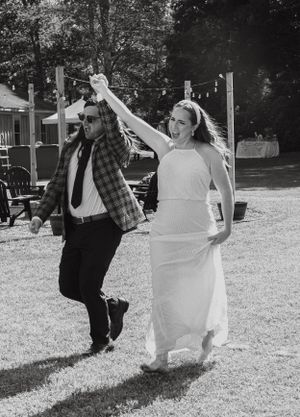 Bride in white dress and groom in plaid jacket dancing joyfully on lawn.