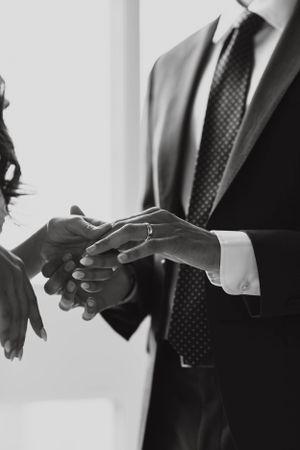 Black and white close-up of groom in a suit placing a wedding ring on the brides hand during ceremony.