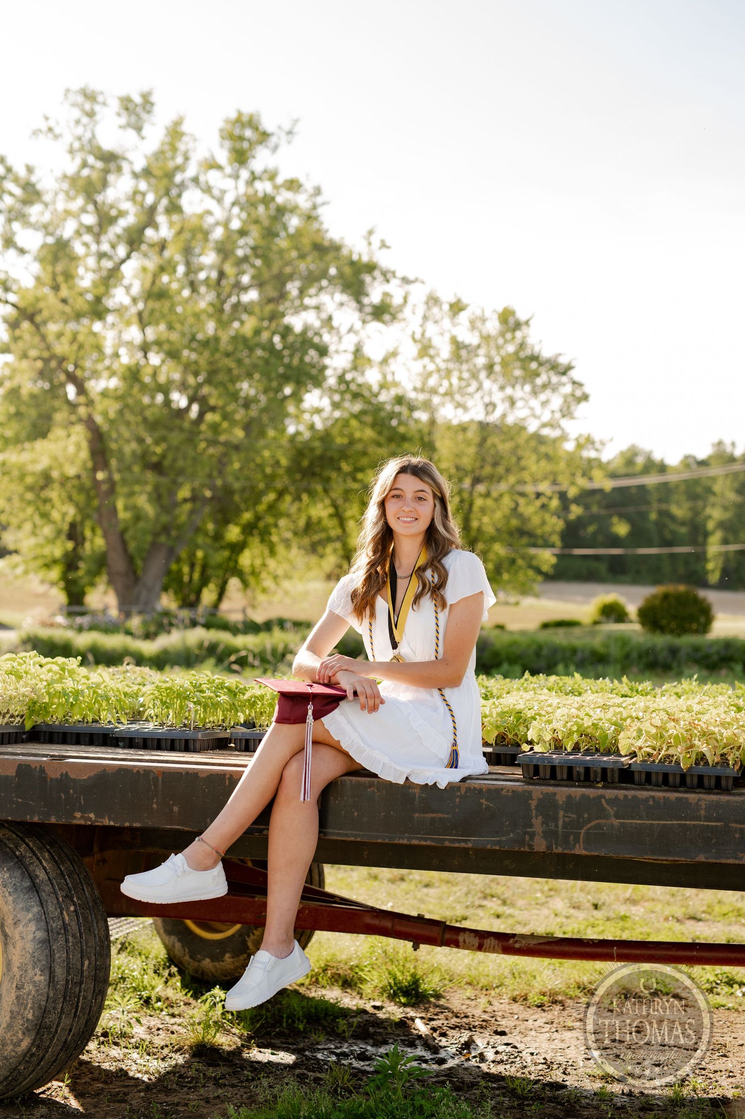 Graduate in white summer dress sits on rustic wagon in sunlit garden setting.