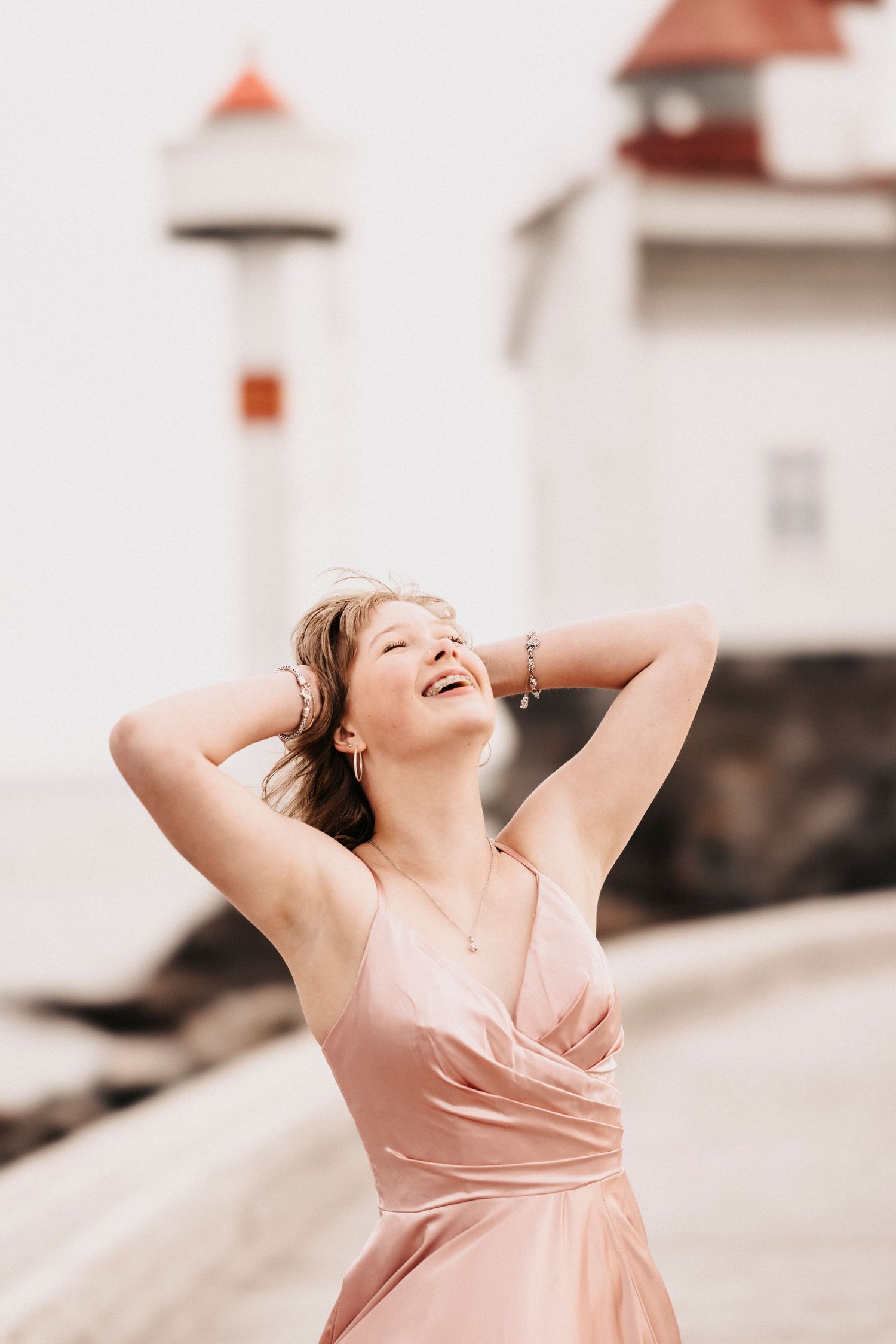 Woman in pink dress laughing joyfully near lighthouse on cloudy day.