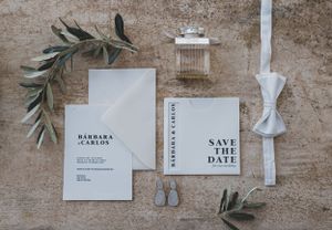 Wedding stationery flat lay with save the date cards, white bow tie, rings and olive branches on textured surface.