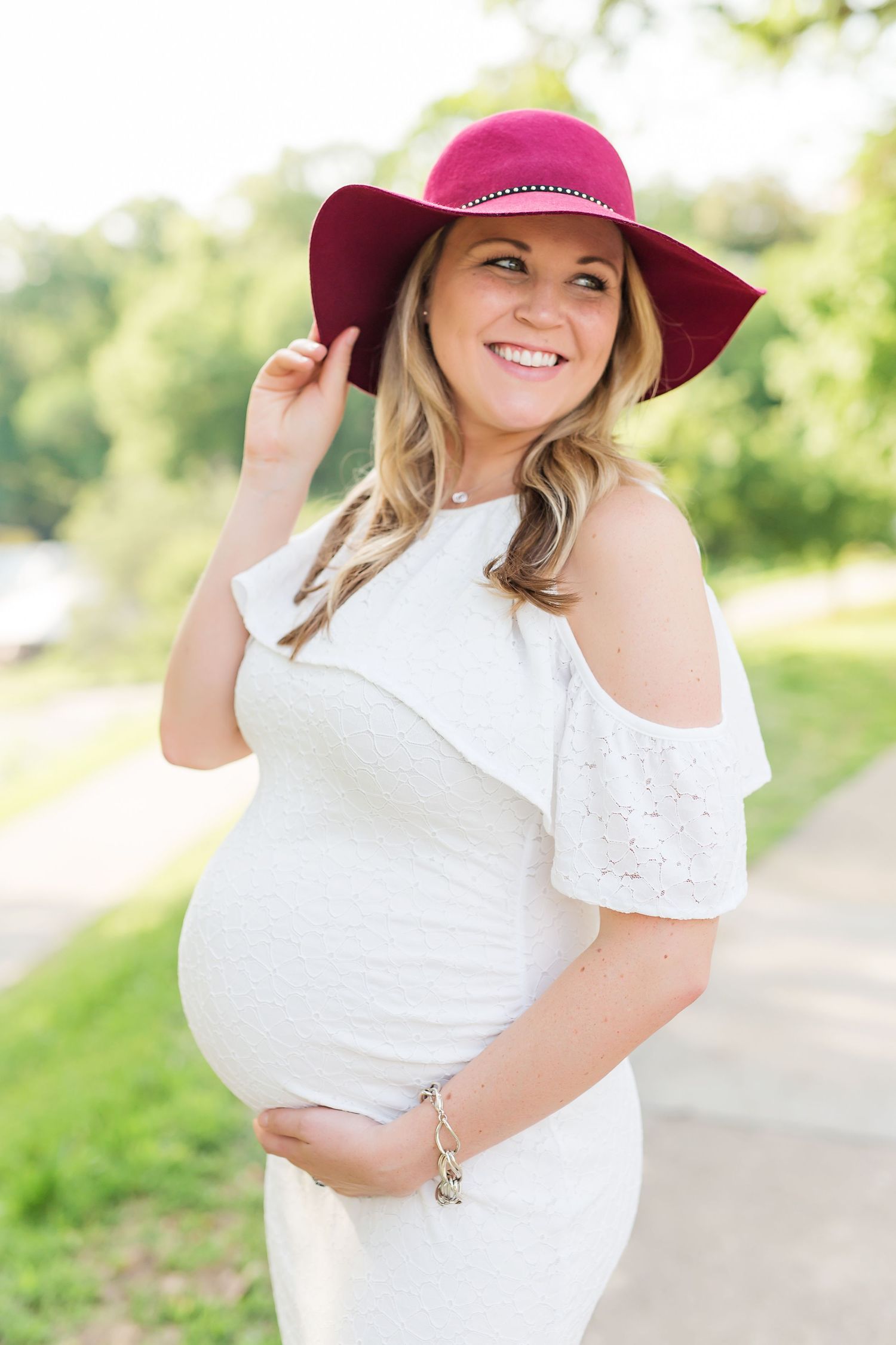 Pregnant woman in white dress and pink sun hat poses outdoors on sunny day.