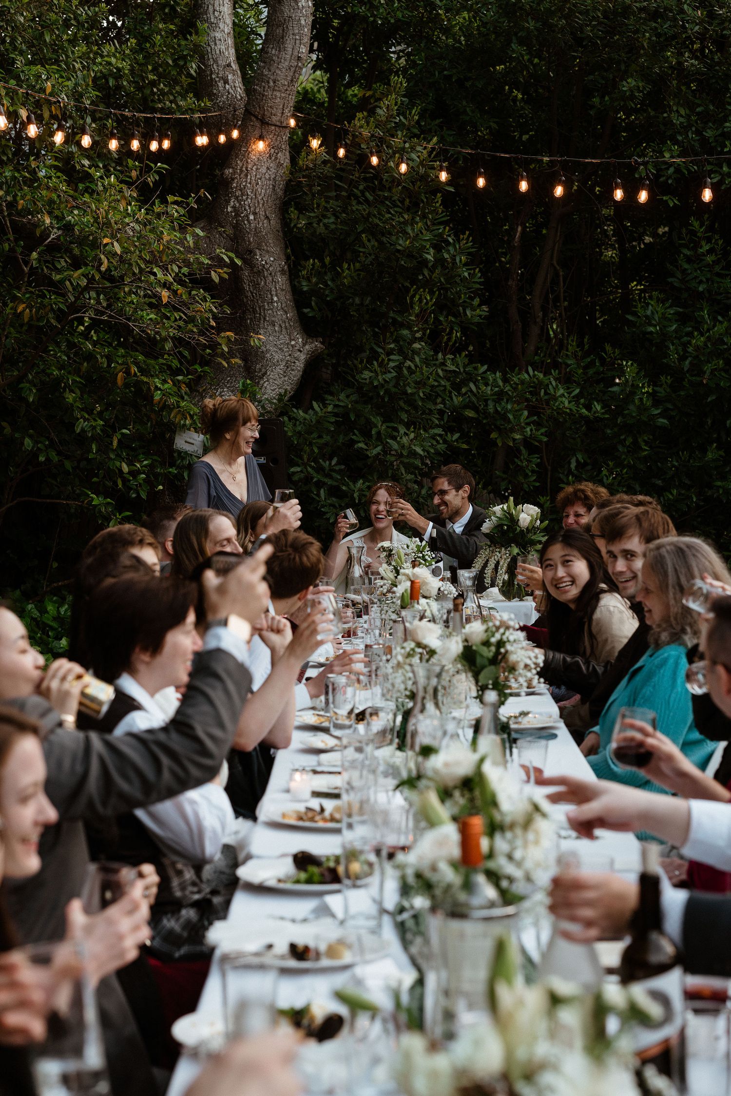 Outdoor wedding reception dinner under string lights with long table of guests.