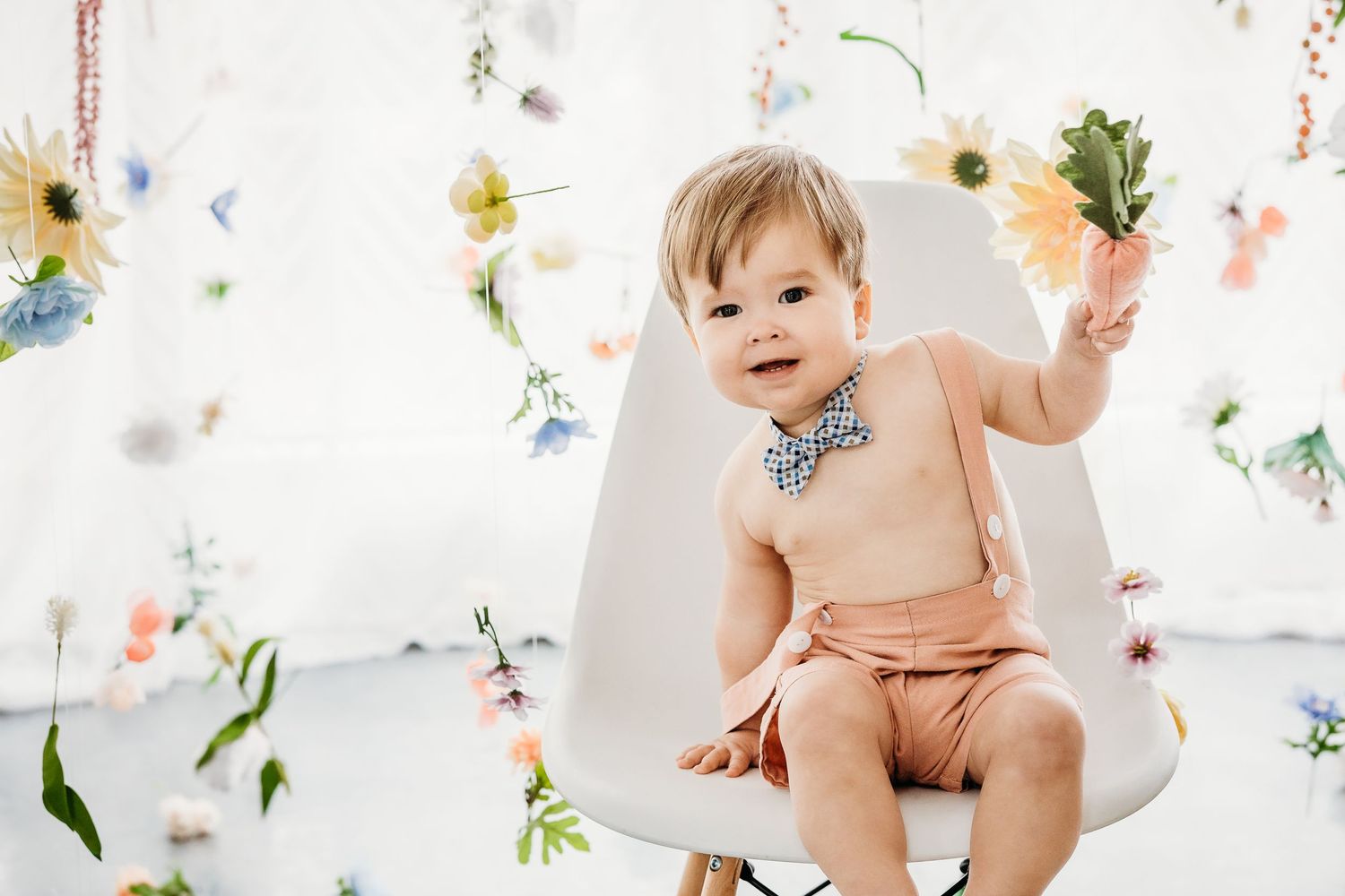 Whimsical first birthday photo with delicate floral decorations and white chair against floral backdrop.