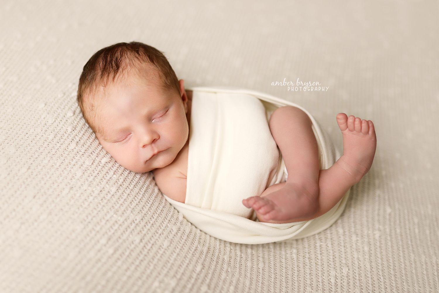 Sleeping newborn wrapped in white fabric lying on textured beige surface.