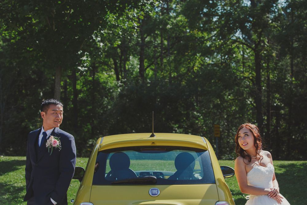 Wedding couple posing beside yellow Fiat car in woodland setting.