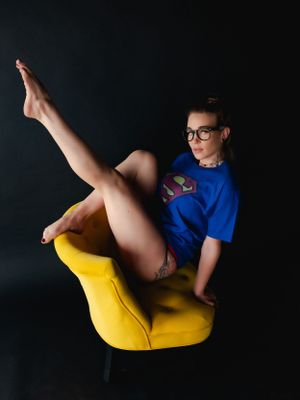 A fangirl in glasses and blue Superman shirt poses for boudoir shoot on a bright yellow modern chair against a dark background.