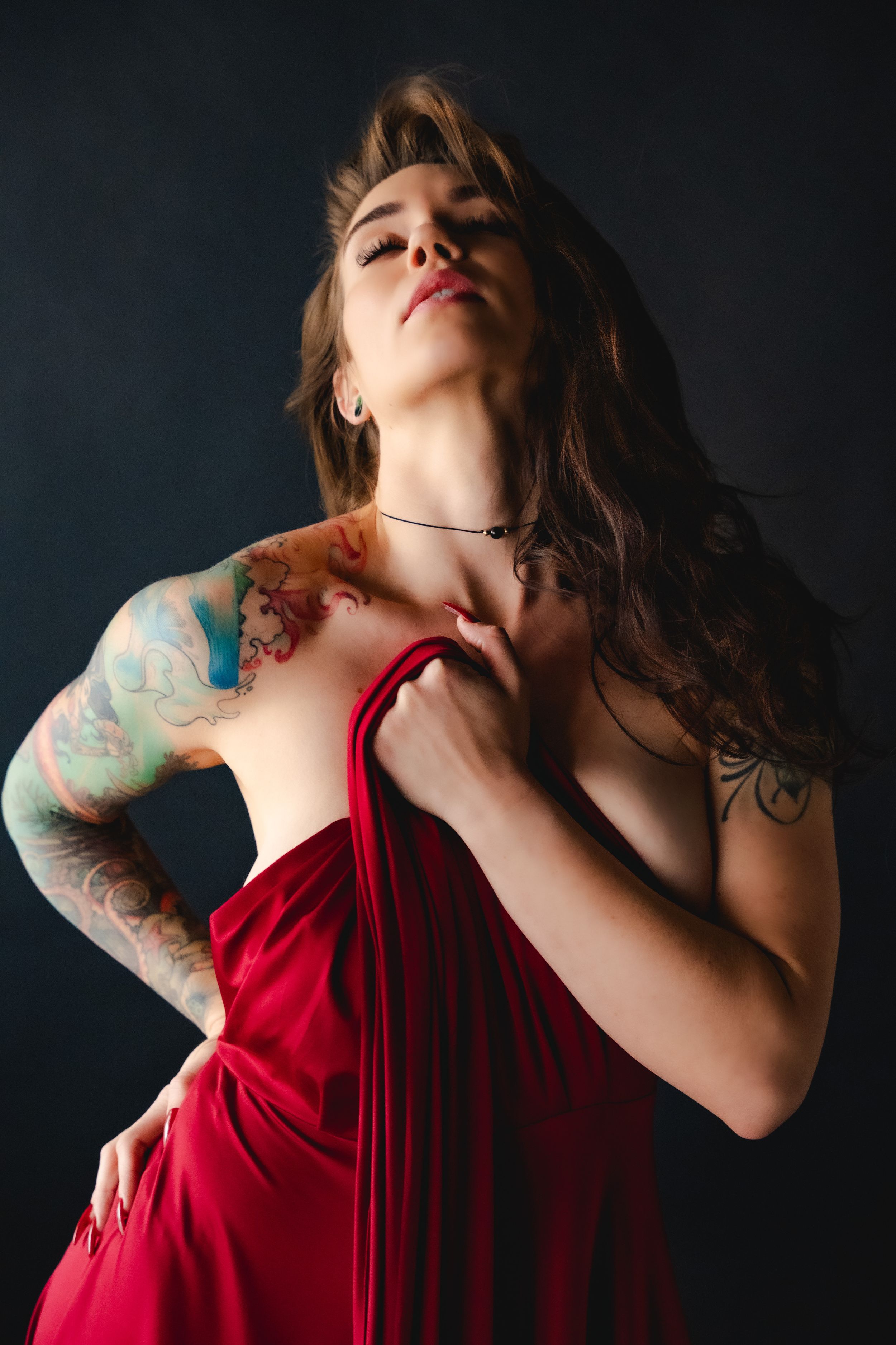 Dramatic boudoir portrait of woman holding up red dress with colorful arm tattoos against dark background in moody lighting.