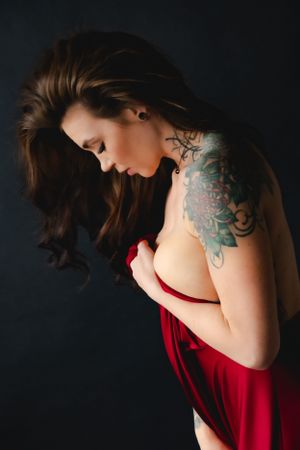 Artistic boudoir portrait of woman in a red dress with long brown hair and colorful shoulder tattoo against dark background.