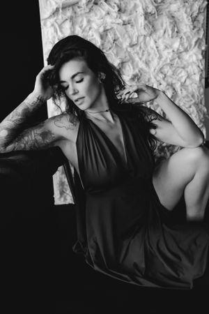 Artistic black and white boudoir portrait of woman in elegant dark halter dress reclining against textured wall with eyes closed.