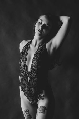 Black and white boudoir photograph showing a woman wearing lace lingerie with arm raised against dark background.