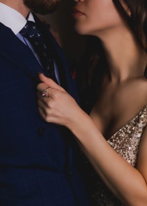 Romantic close-up boudoir image of couple in formal wear showing engagement ring, with navy suit and sequined dress details.