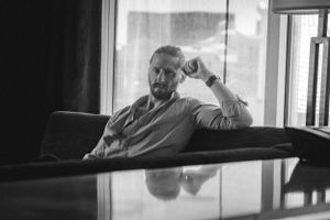 A male boudoir image of him resting casually on a couch by a window with a reflection in a black and white photograph.