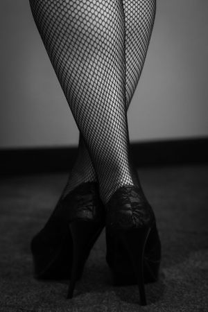Black and white photo of crossed legs in fishnet stockings and high heel shoes against dark background.