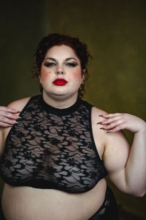 Boudoir portrait of plus sized woman in black lace halter top against dark background showcases dramatic makeup and vintage styling.