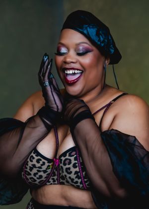 A joyful boudoir portrait of a plus sized black woman in black gloves and hat posing against a dark background.