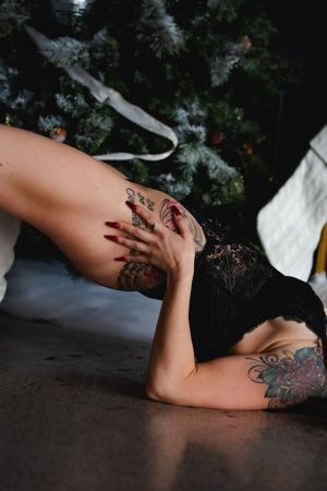 Artistic boudoir photo showing intricate tattoos on a person posing in black lingerie against a dark background and Christmas tree.