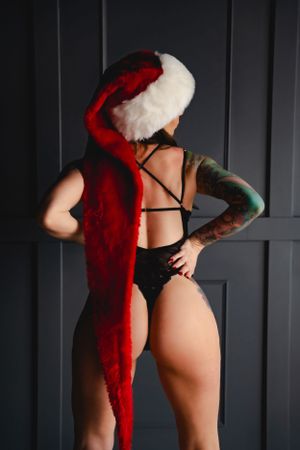 Woman wearing Santa hat and poses in black lingerie against dark wall showing artistic holiday boudoir style.