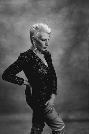 Black and white portrait of an older woman in sequined top and light pants striking dramatic pose against moody backdrop.