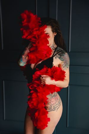Artistic boudoir photo featuring bright red feather boa and visible tattoo artwork against dark background.