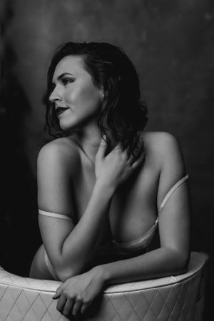 Artistic black and white boudoir portrait of a woman in lingerie posing on quilted furniture against dark backdrop.