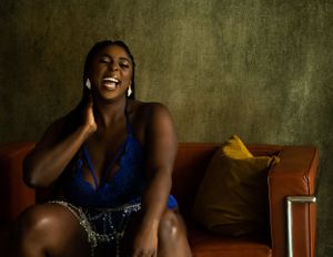 A joyful black woman in a blue lingerie laughs while sitting on a brown leather couch against a textured olive backdrop.