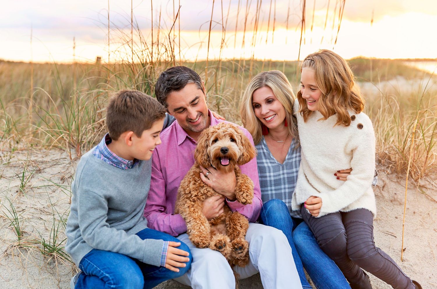 Family and dog relaxing together on a beach at sunset among sea oats and dunes.