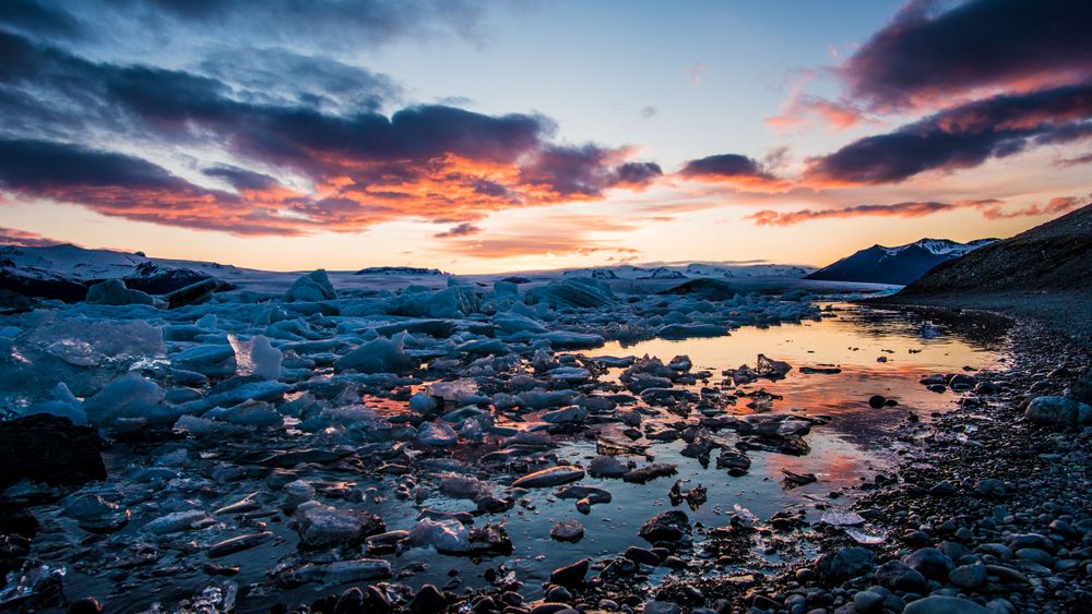 Ice-covered rocky shore reflects vibrant sunset colors across frozen landscape.