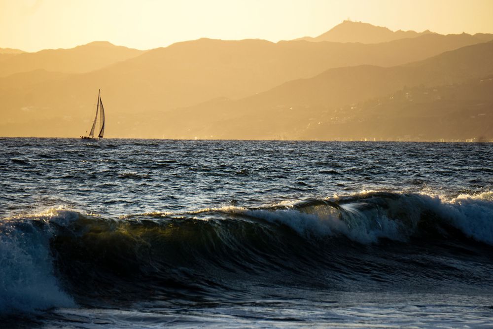 A sailboat silhouetted against mountains as waves crash during golden hour.