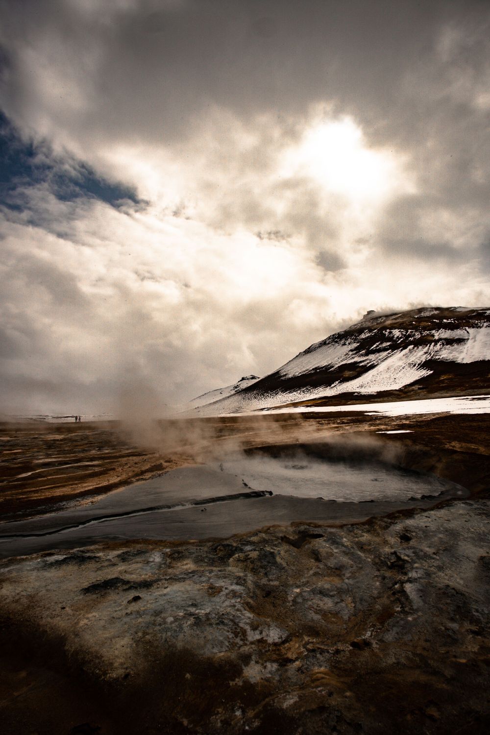 Steam rises from a geothermal pool against a dramatic cloudy sky and snow-capped mountain.