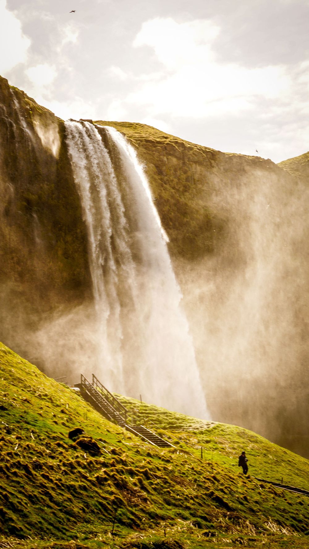 Massive waterfall cascades down green mountainside with wooden walkway and tiny figure in distance.