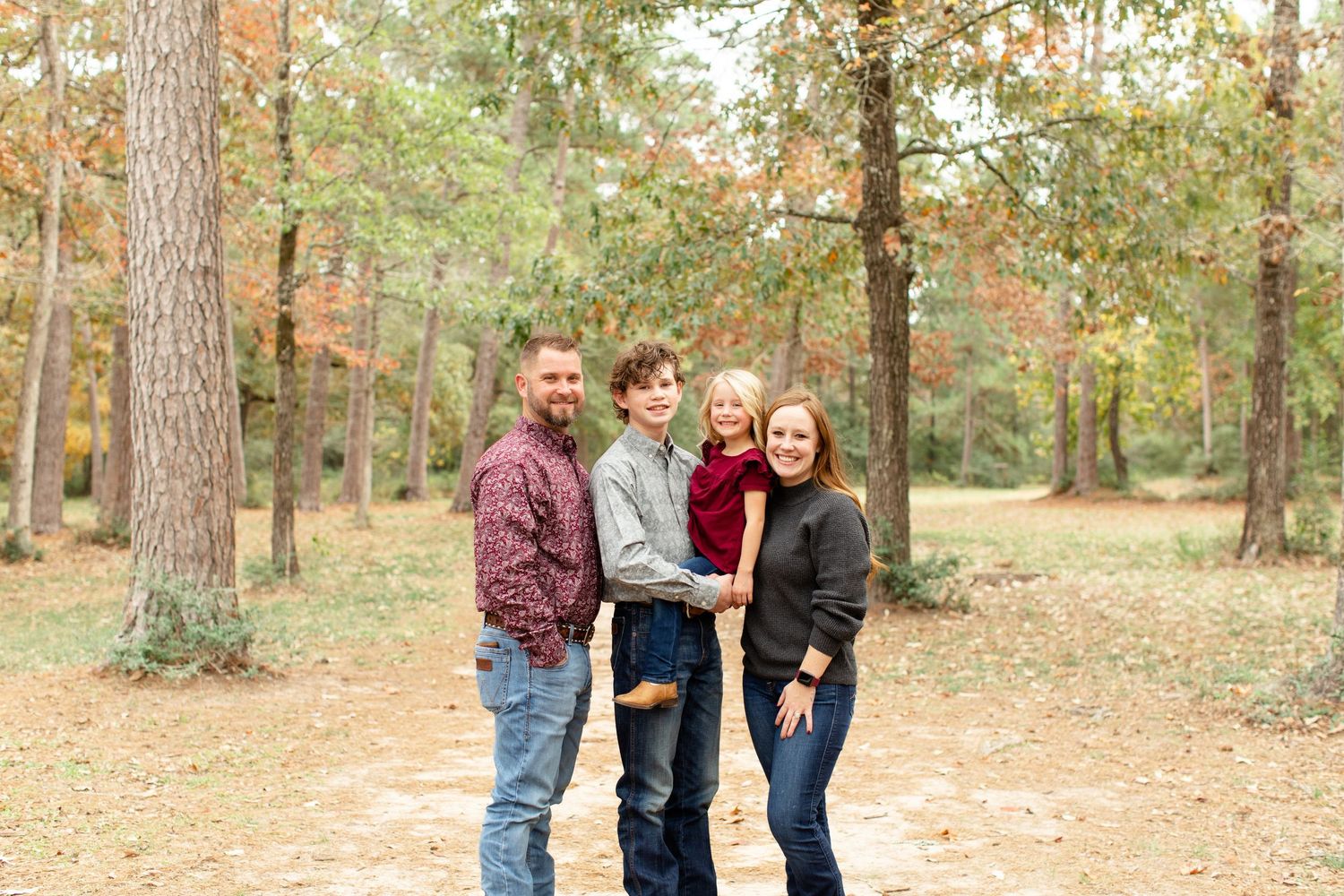 A family stands together on a forest trail during autumn with pine trees in the background.