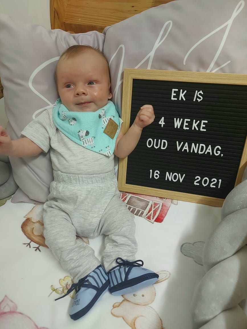 Baby in grey outfit lying next to letter board displaying date and age milestone.