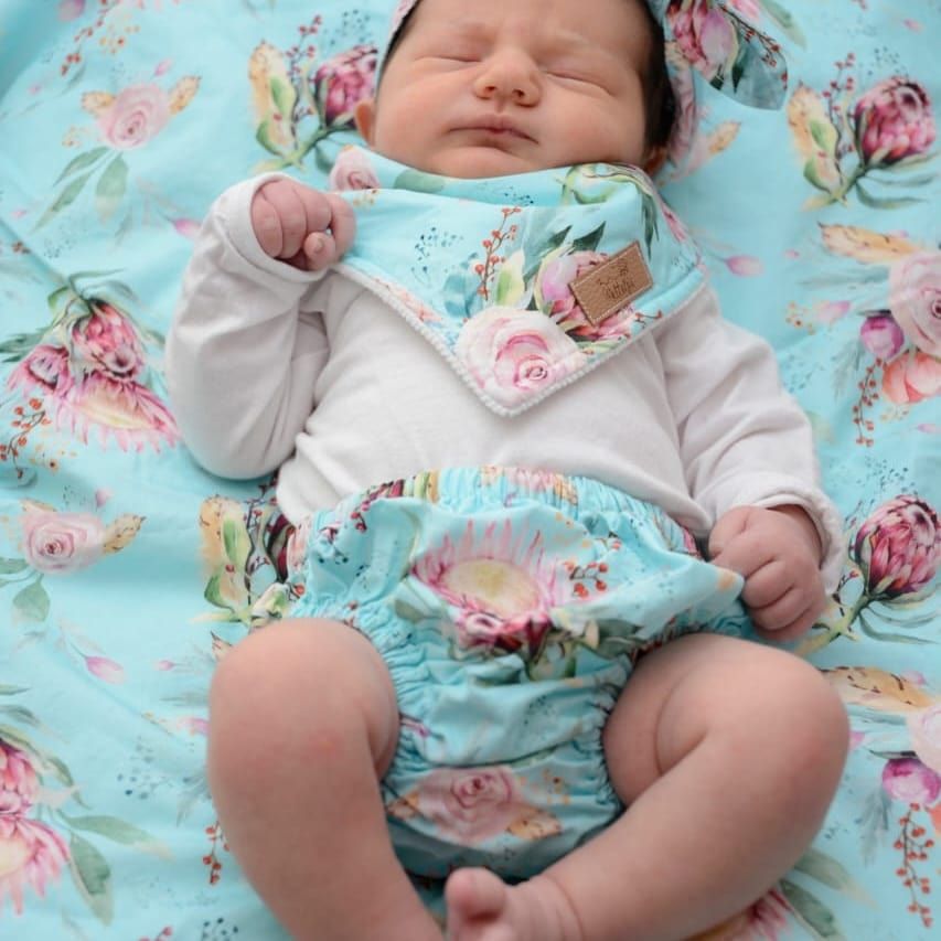 Baby wearing white onesie resting on blue floral bedding.