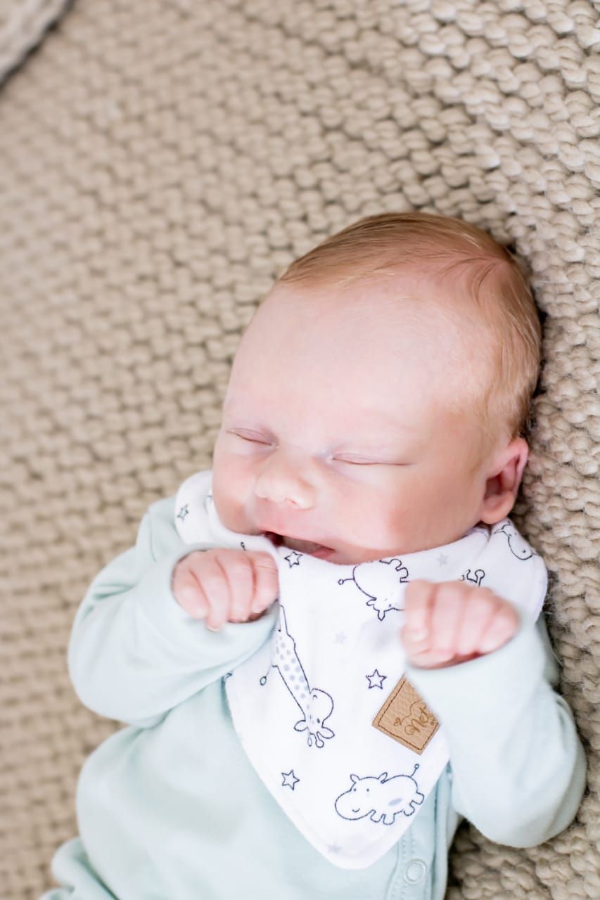 Baby wearing white bib with animal print design rests on textured beige surface.