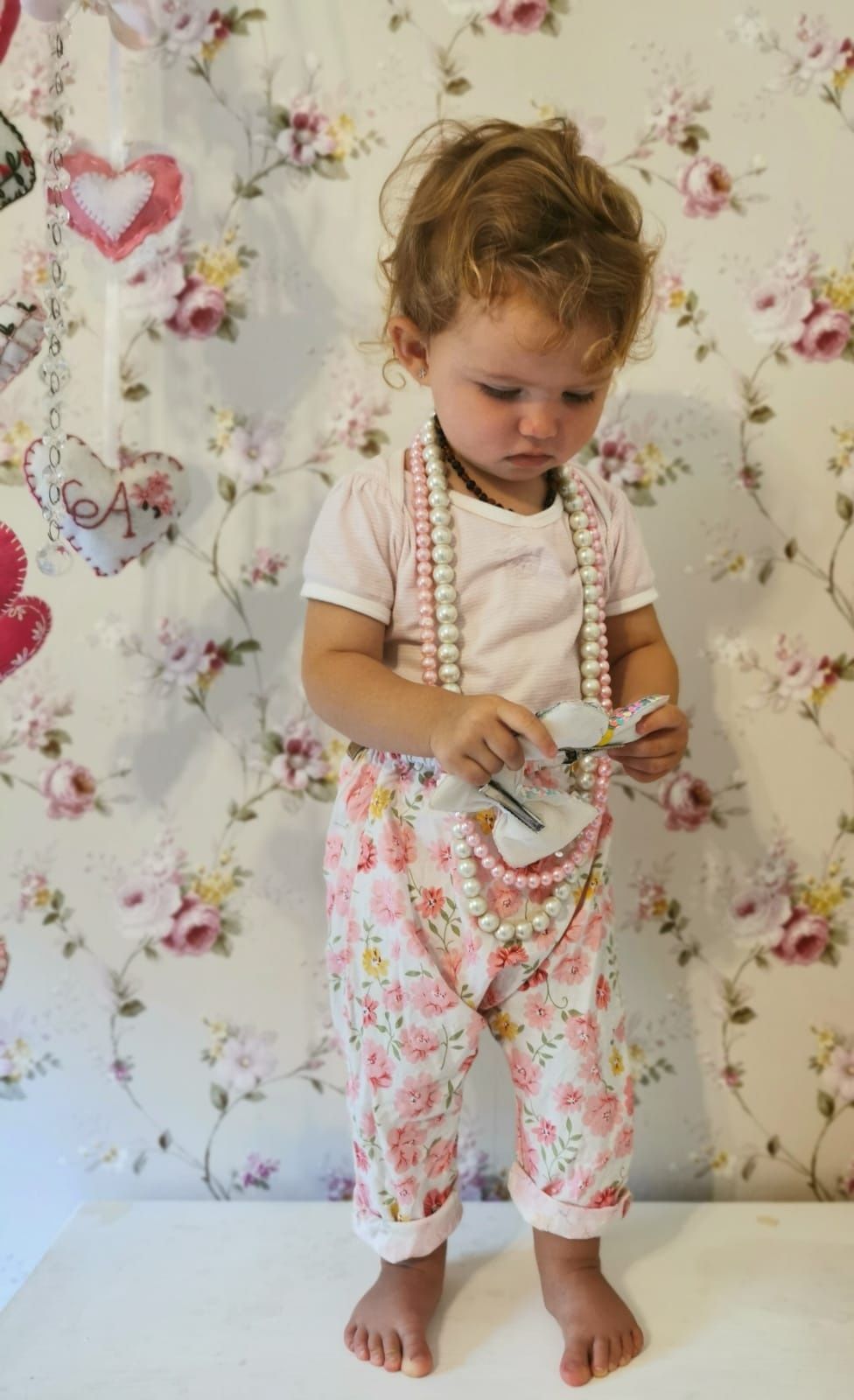 Toddler in floral outfit with pearl accessories playing against floral wallpaper background.