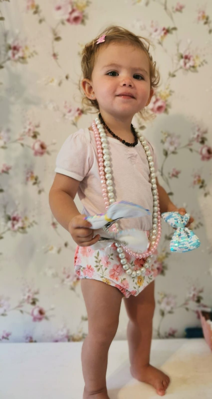 Small child wearing vintage-inspired outfit with pearl accessories against floral wallpaper.