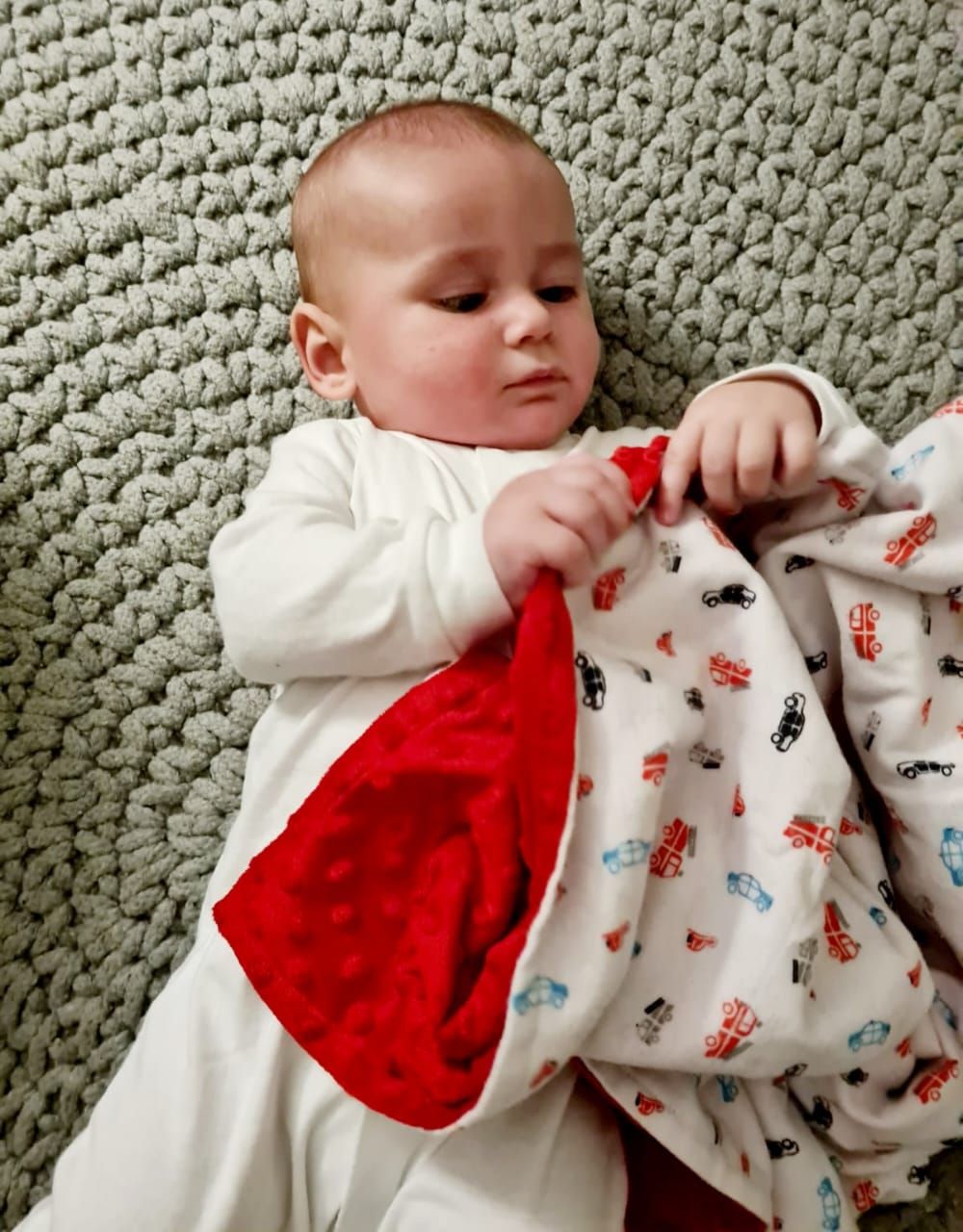 Baby in white outfit playing with red minky blanket on textured green surface.