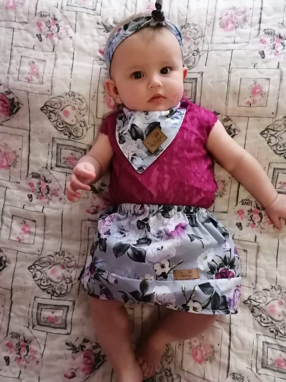 Baby wearing a pink and gray patterned dress lies on floral patterned fabric.