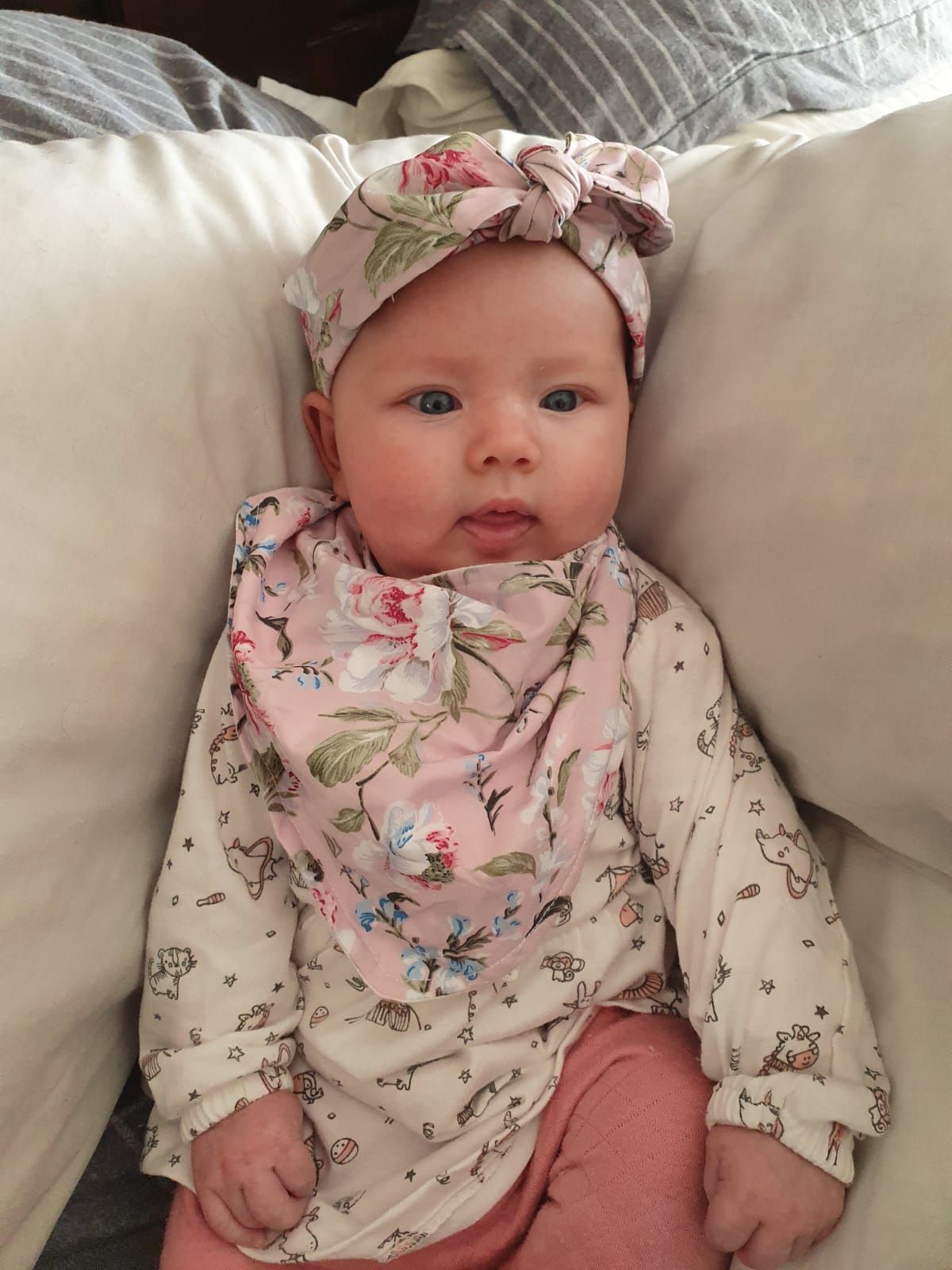 Baby wearing floral print outfit with matching headband rests on cream cushions.
