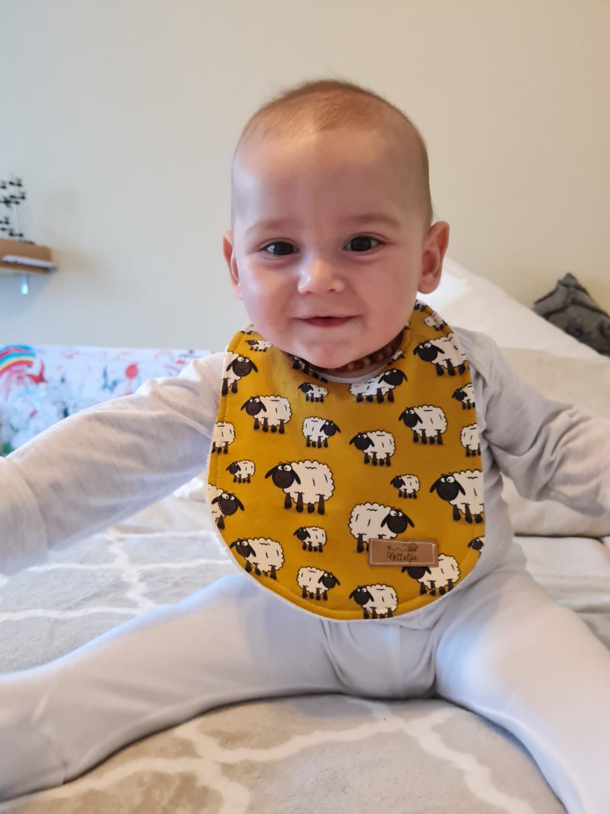 Baby wearing white outfit with yellow sheep-patterned bib lying on white bedding.