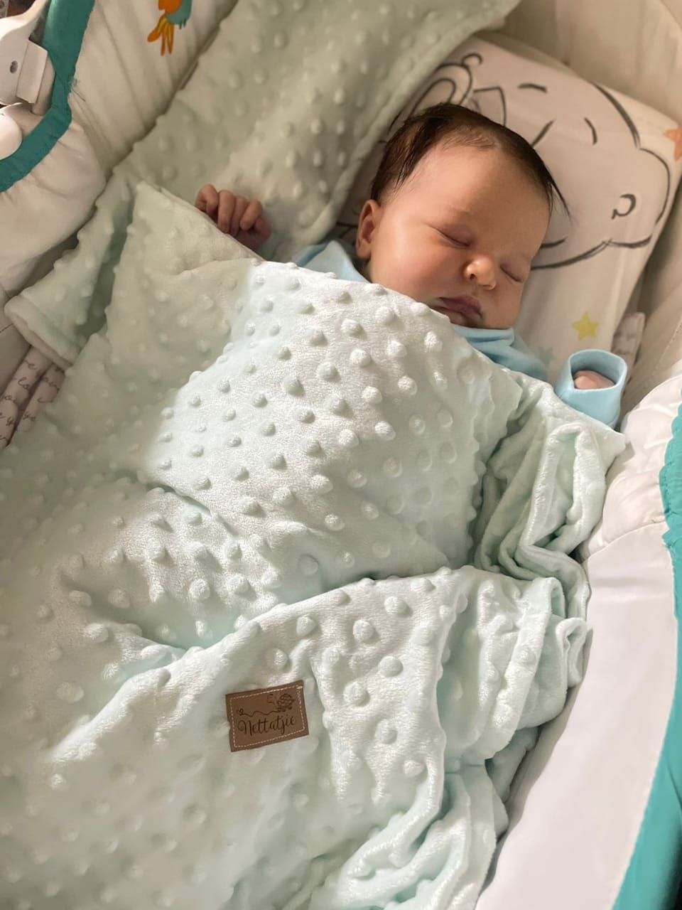 Baby sleeping under mint green dotted blanket on white bedding.