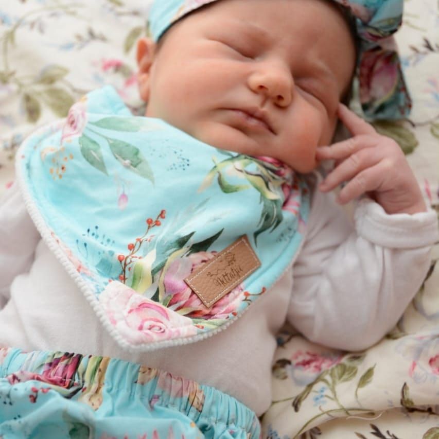 Baby wearing mint green bib rests on floral patterned bedding.