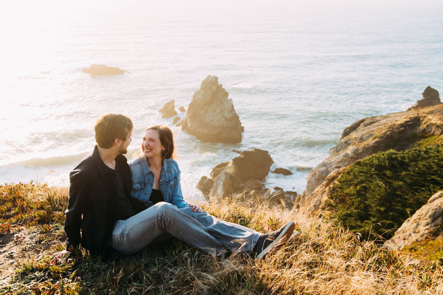 Couple sits in grassy area at Lands End in San Francisco for engagement portraits.