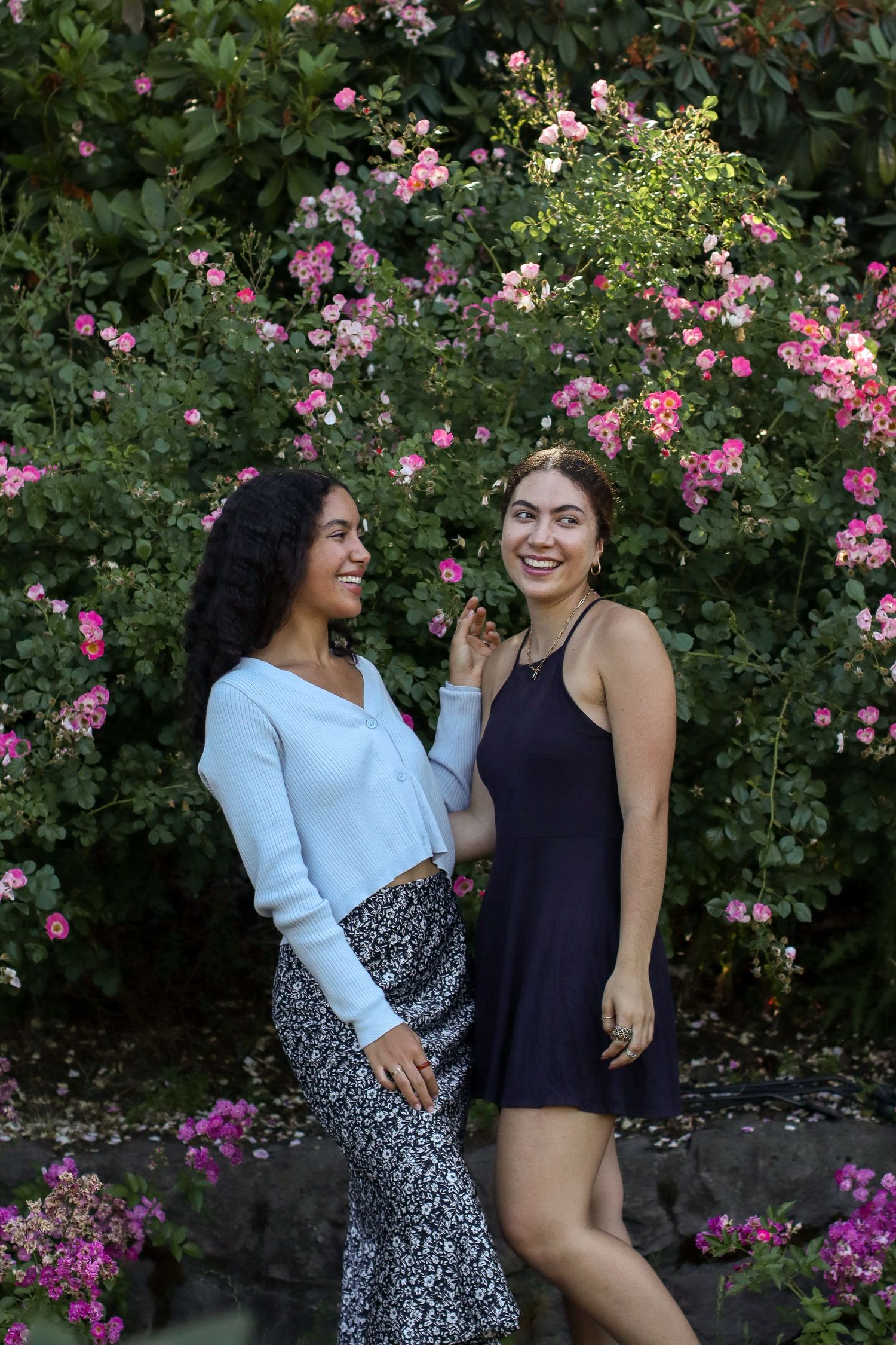 Two friends share laughter together in front of blooming pink rose garden.