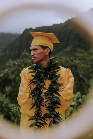 Graduate in yellow cap and gown with green lei stands against mountain backdrop.