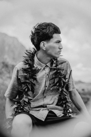 Person wearing grey shirt and dark lei poses against cloudy mountain backdrop.