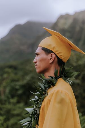 Graduate in bright yellow regalia and dark lei gazes at scenic mountain landscape.