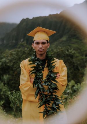 Yellow-robed graduate wearing traditional green lei poses outdoors with mountains in background.